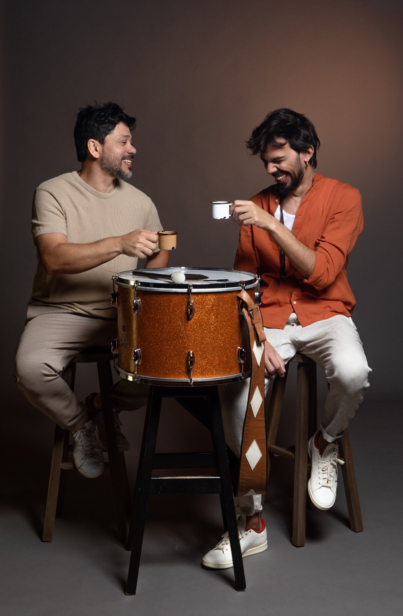 Guegué Medeiros e Salomão Soares rindo e brindando com canecas de café, sentados em volta de uma zabumba